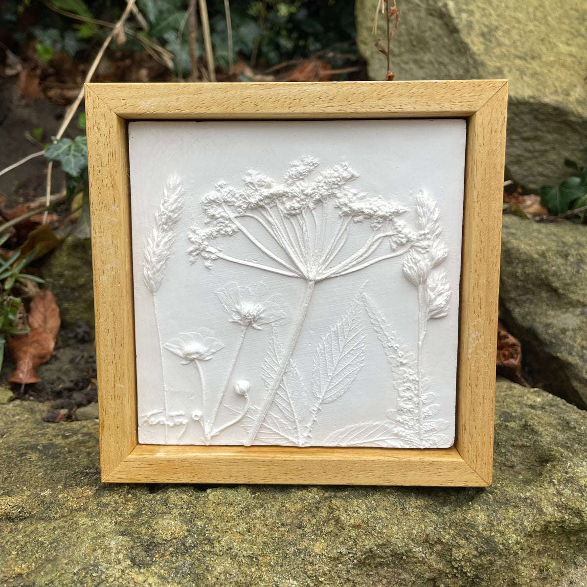 A square framed wall art piece featuring a white embossed floral and botanical design, set in a light natural wood frame and photographed outdoors on a stone surface with foliage and rocks in the background.