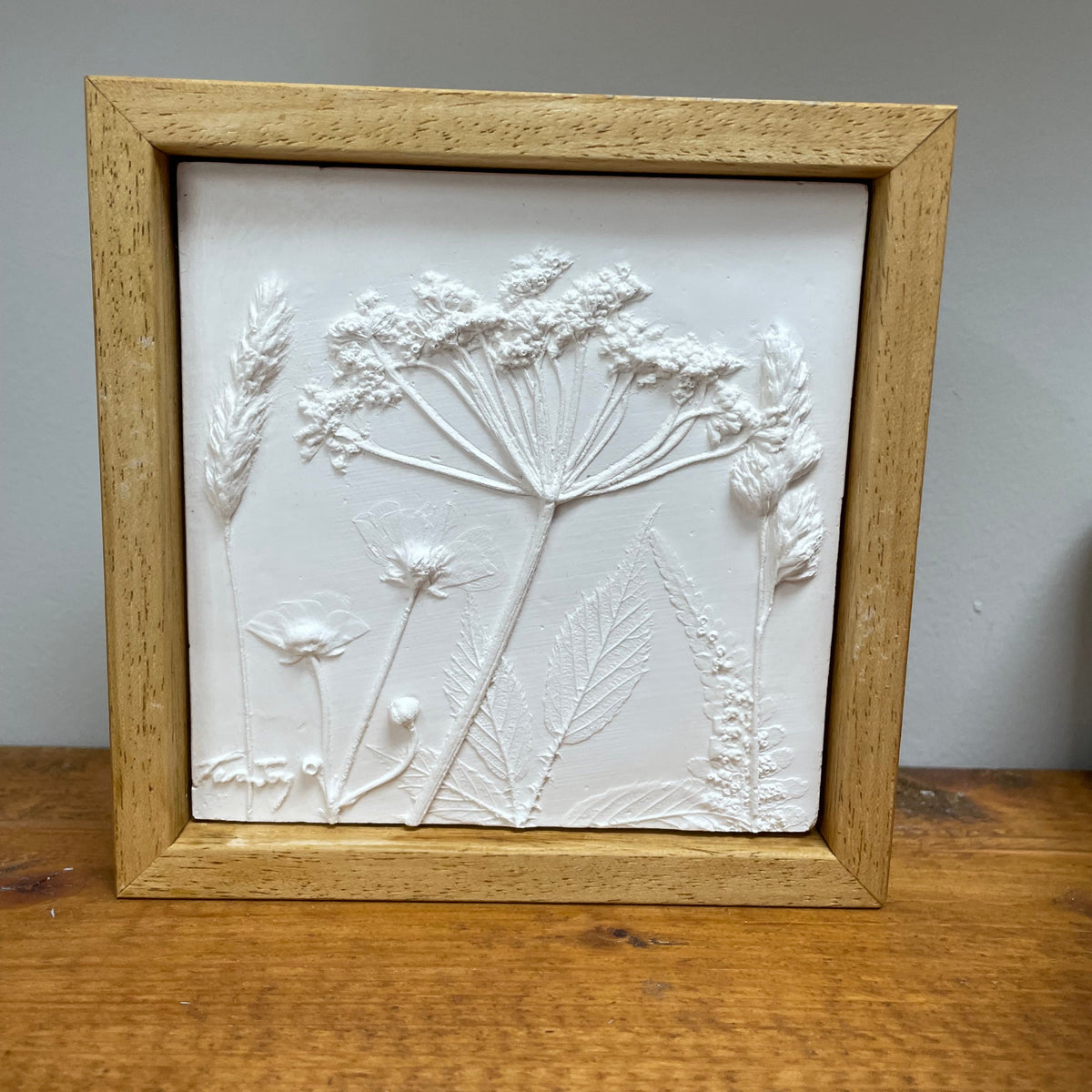 A square framed white relief wall plaque featuring embossed botanical and wildflower designs, set in a light natural wood frame and displayed on a wooden surface against a gray wall.