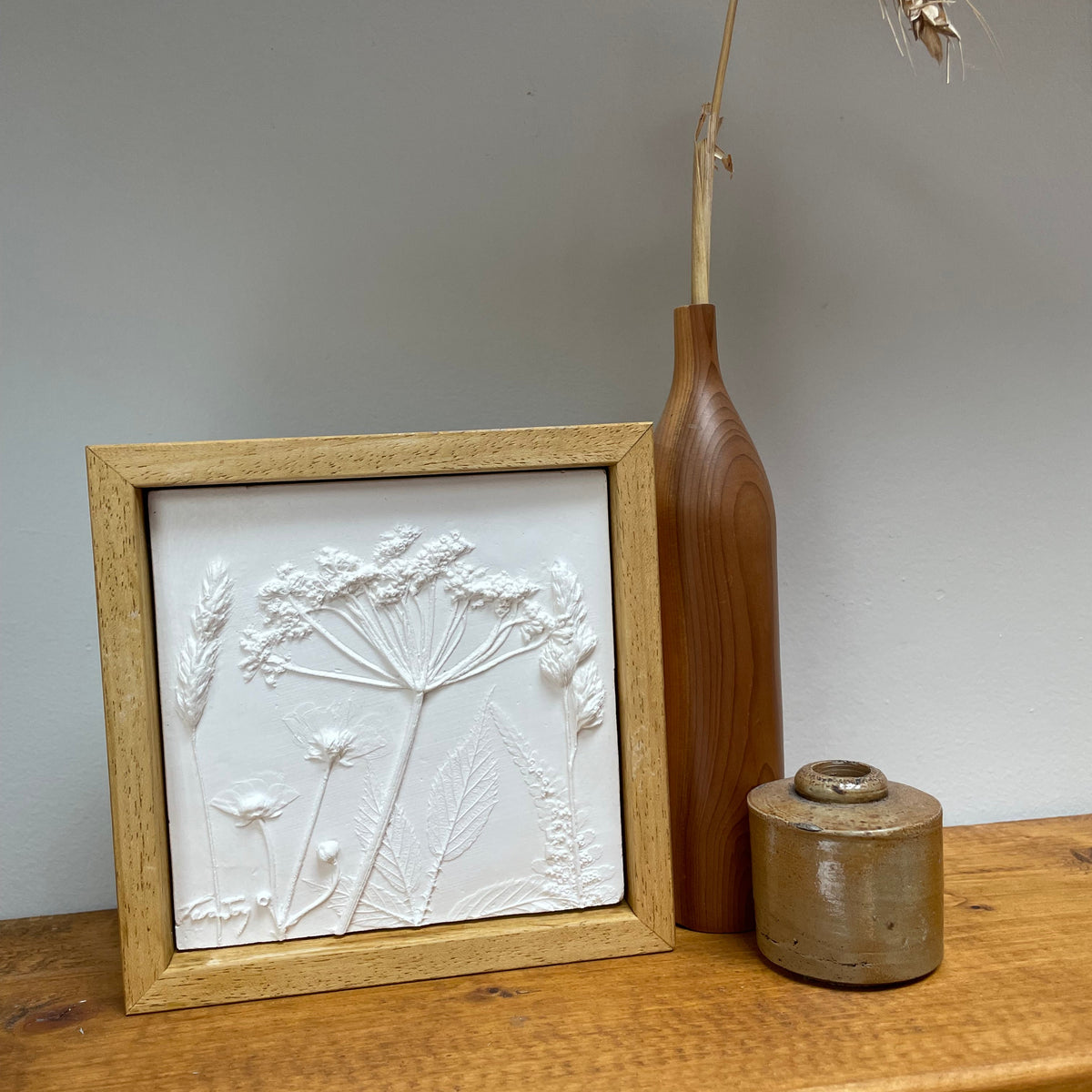 A square framed white relief wall art piece with embossed wildflower stems, displayed on a wooden shelf beside a tall brown vase with dried wheat and a small ceramic jar against a plain light gray wall.