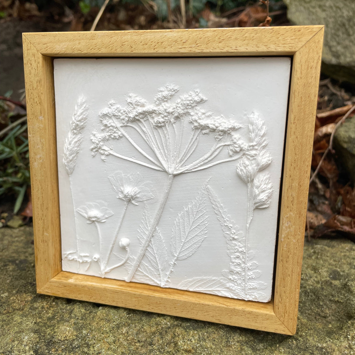 A square framed white relief wall plaque featuring embossed wildflower botanical designs, set in a light natural wood frame and photographed outdoors on a stone surface with foliage in the background.