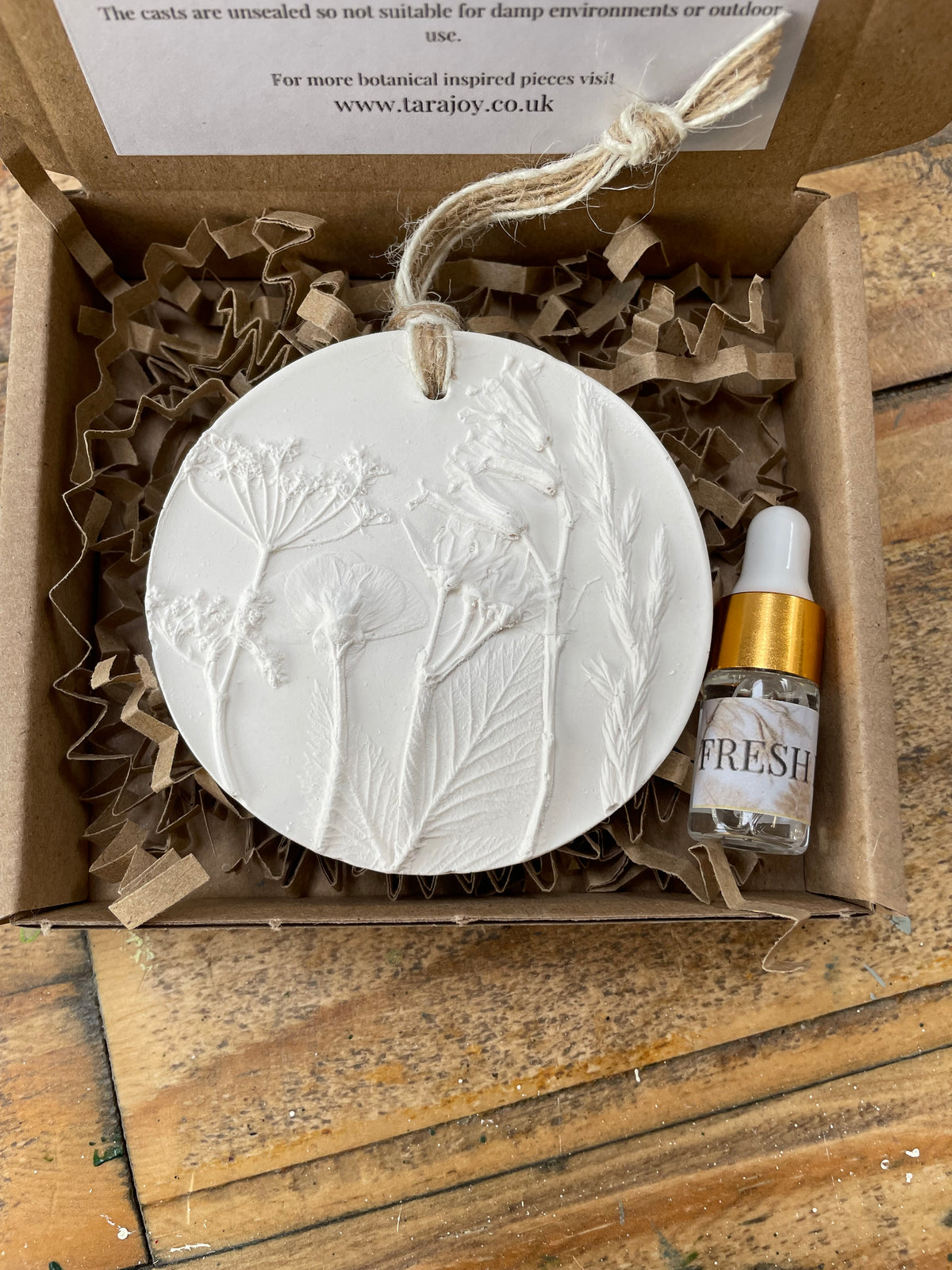 A round white sculpture plaster botanical ornament with embossed leaves and flowers, displayed in an open cardboard gift box with crinkle paper filler and a small glass dropper bottle labelled FRESH Linen. A product card is visible inside the box.
