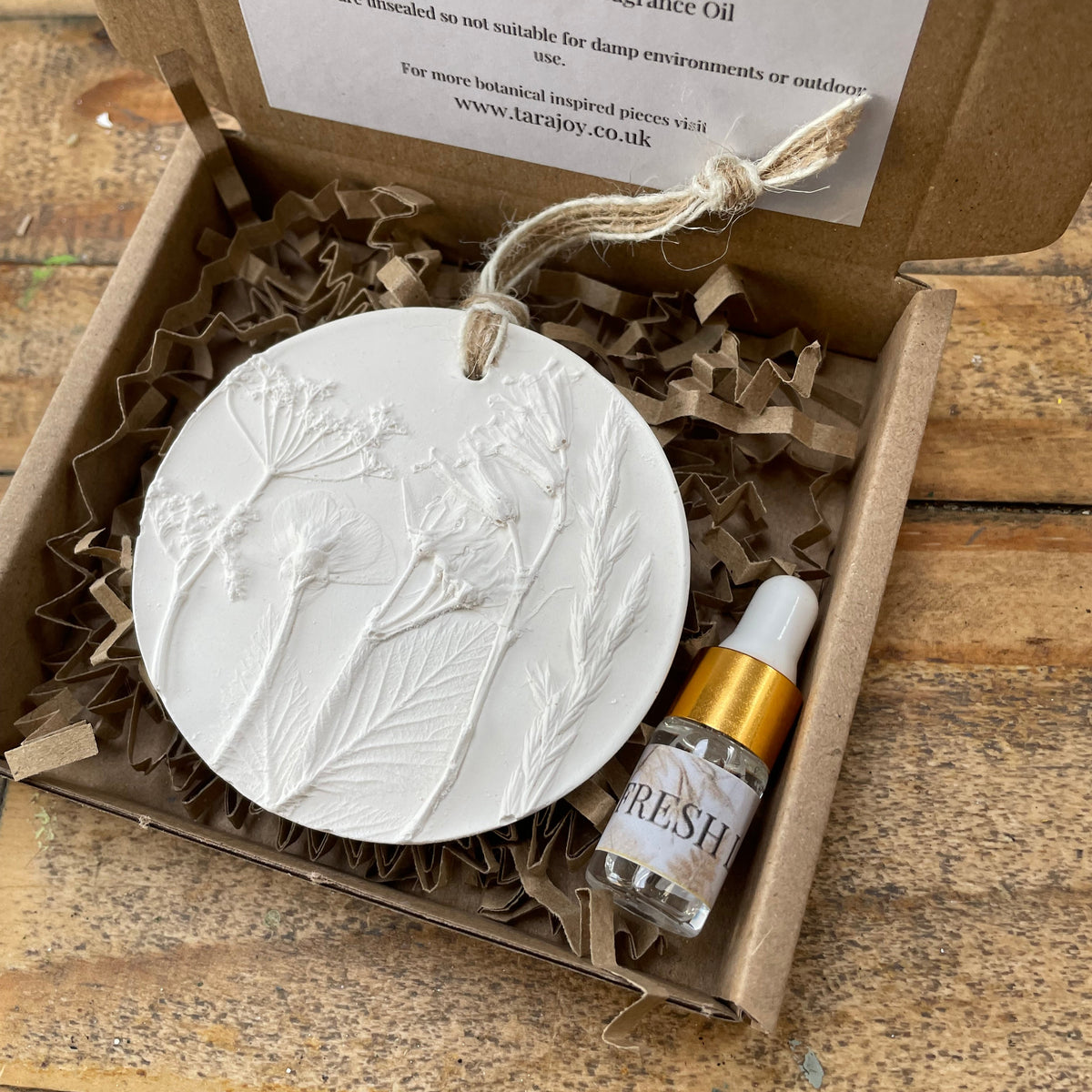 A gift box containing a white carved sculpture plaster round ornament with a hanging loop, a small glass dropper bottle labeled 'Fresh Linen' with a gold cap, and a printed card, arranged on a rustic wooden surface.