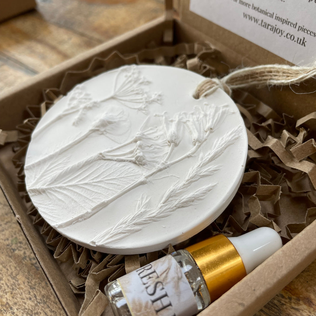 A white oval carved sculpture plaster ornament with floral and leaf engravings, displayed in a gift box next to a small clear bottle with a white cap and gold collar. The box contains brown crinkle paper packing material.