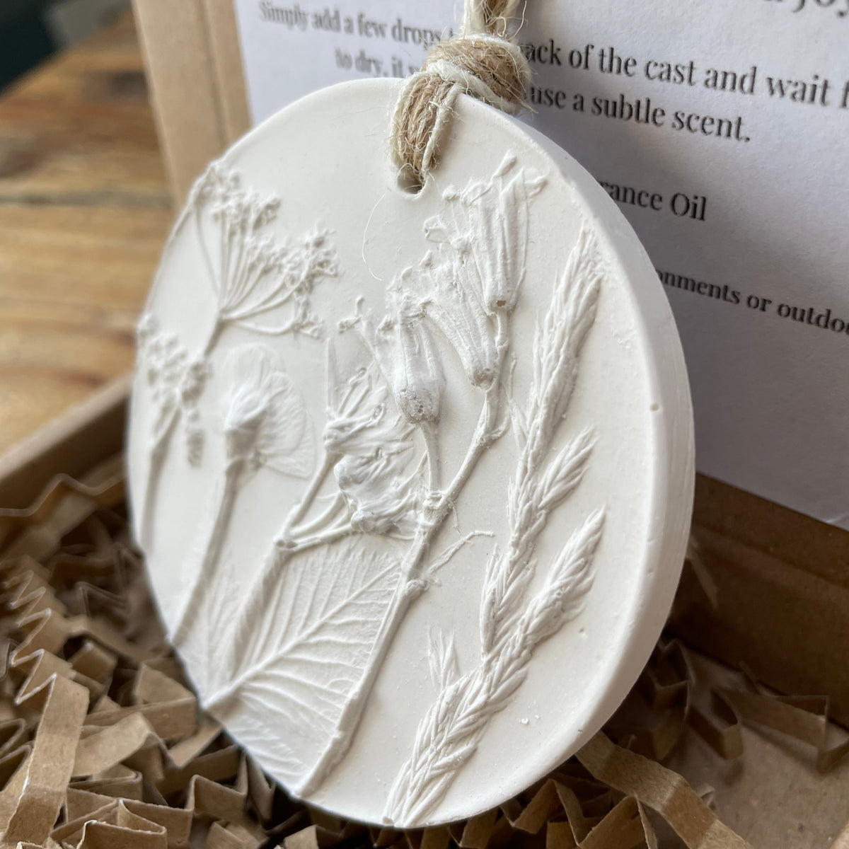 A round off-white sculpture plaster ornament featuring embossed wildflower and botanical designs, hanging from a natural jute twine loop. The ornament is displayed in brown paper shred inside a gift box, with a printed card partially visible in the background.
