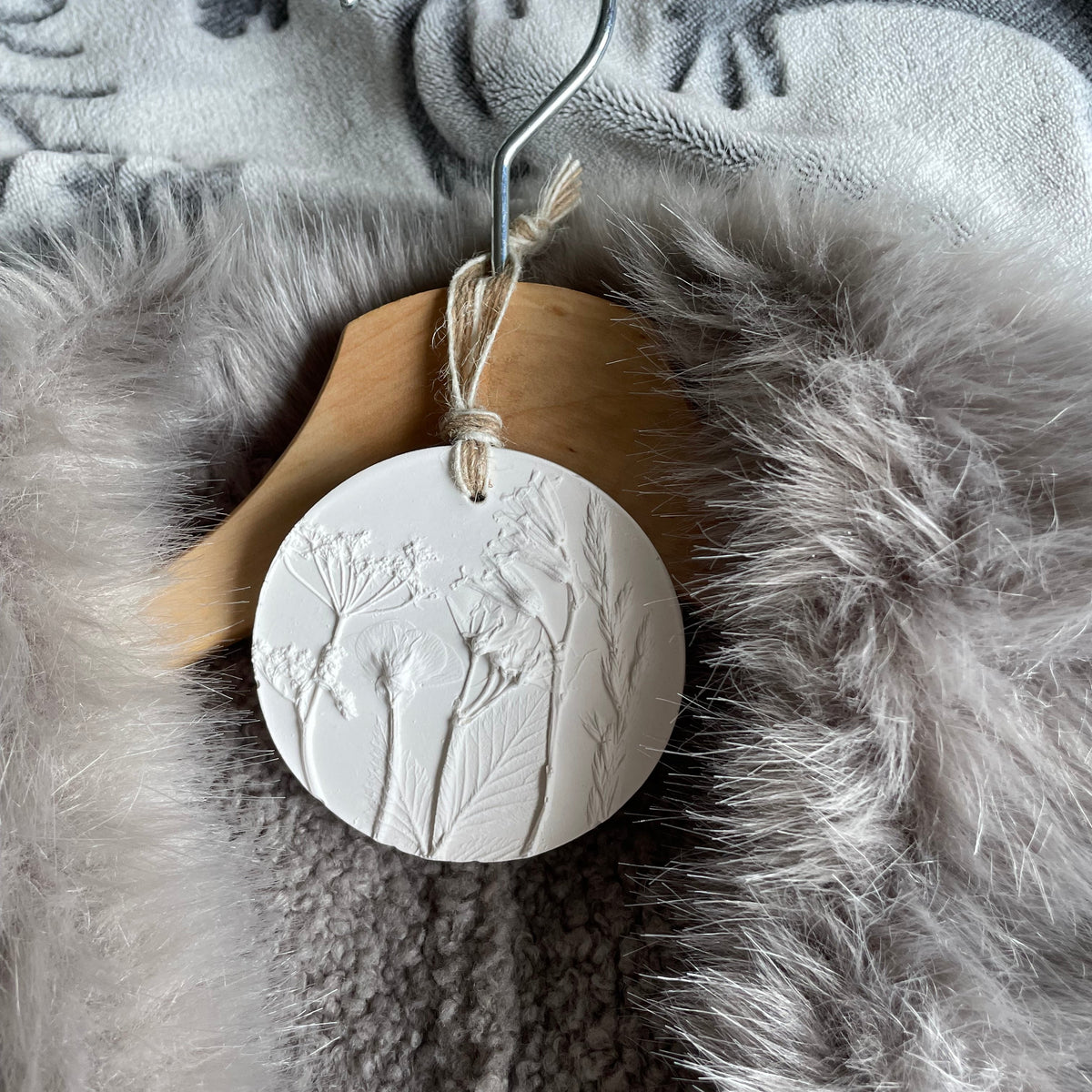 A round white sculpture plaster ornament with embossed wildflower details hangs from a wooden hanger with a metal hook, set against a grey faux fur and textured blanket background.