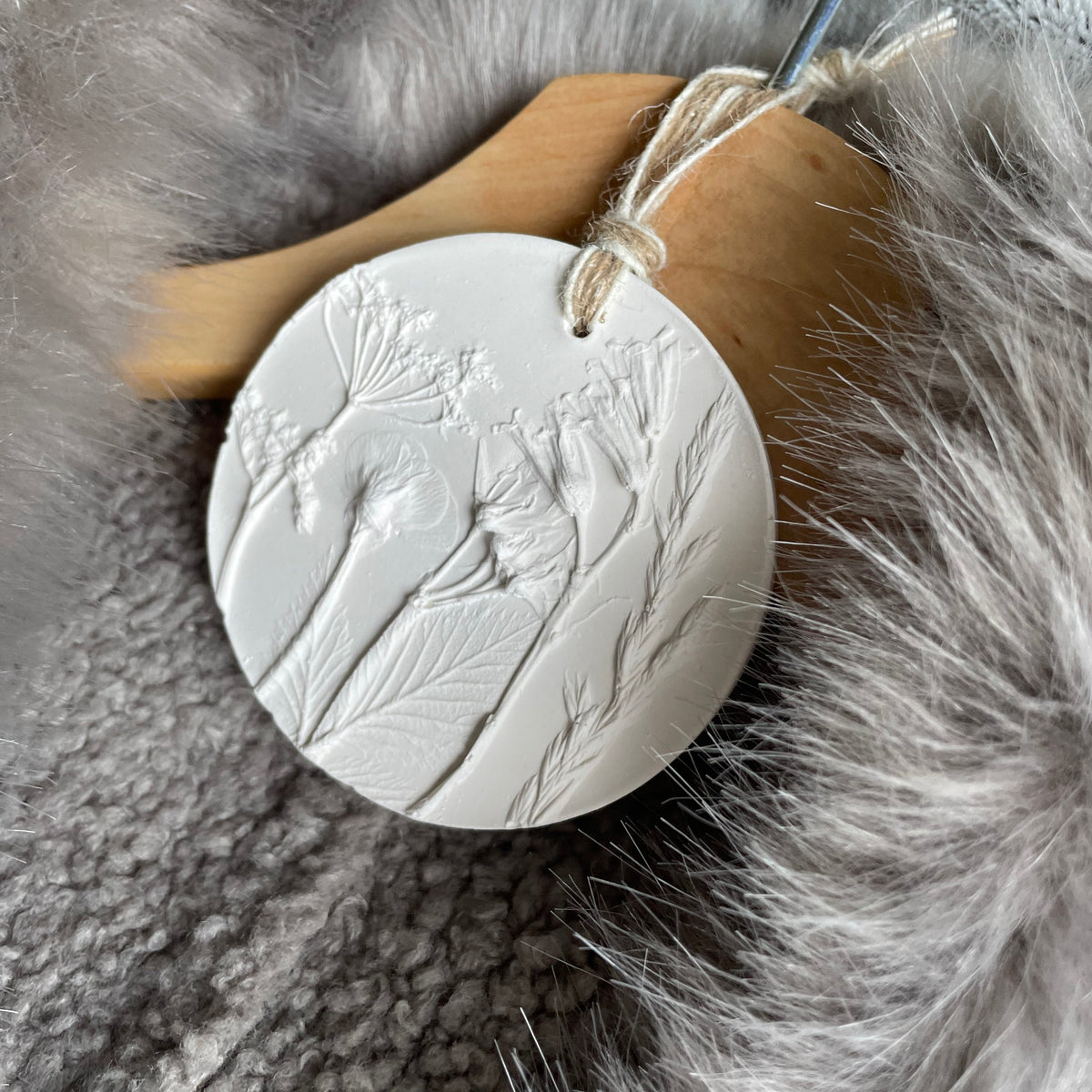 A round white sculpture plaster ornament with embossed wildflower details, hanging from a wooden hanger with a metal hook, set against a gray faux fur background.
