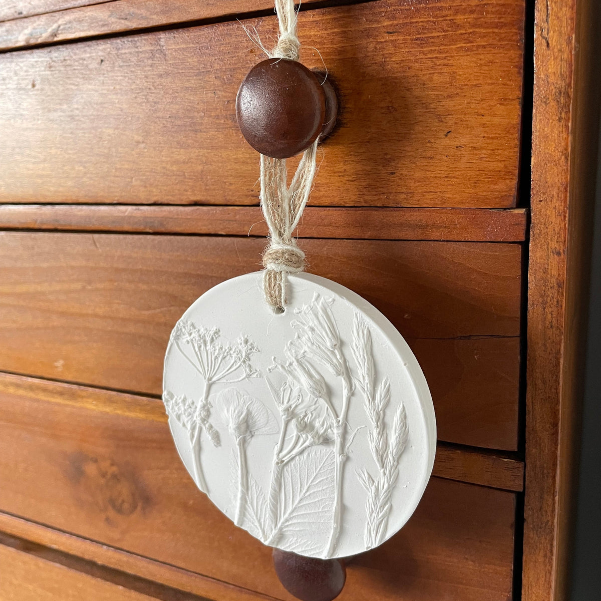 A round white sculpture plaster ornament featuring embossed wildflower designs, hanging from a wooden drawer knob on a rustic wooden chest.